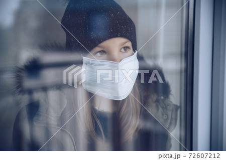 A girl in a black cap with a protective mask stands by the window. 72607212