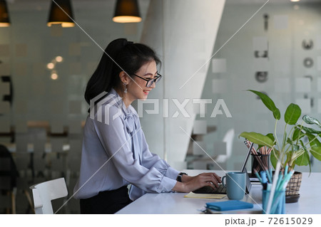 Side view of cheerful young woman sitting in front of her work desk and working with computer tablet. 72615029