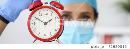 Doctor in medical protective mask holds red alarm clock in his hands Doctor in medical protective mask holds red alarm clock in his hands 72620016