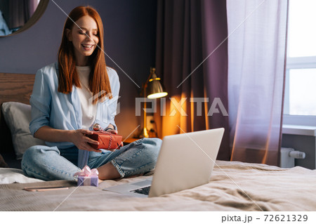 Cheerful young woman holding gift box with present during online communication using laptop. Cheerful young woman holding gift box with present during online communication using laptop. 72621329