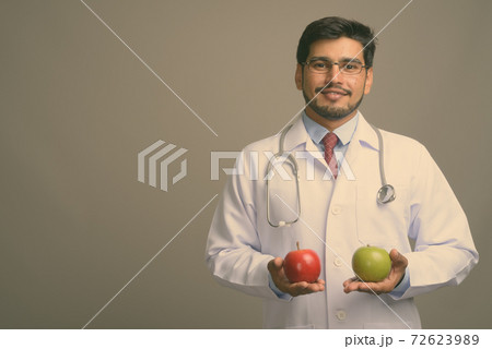 Young handsome bearded Persian man doctor against gray background Young handsome bearded Persian man doctor against gray background 72623989