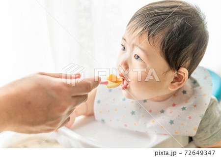 窓辺でベビーチェアに座って離乳食を食べる赤ちゃん 72649347