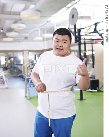 asian overweight man measuring waist and happy with progress in gym asian overweight man measuring waist and happy with progress in gym 72654572