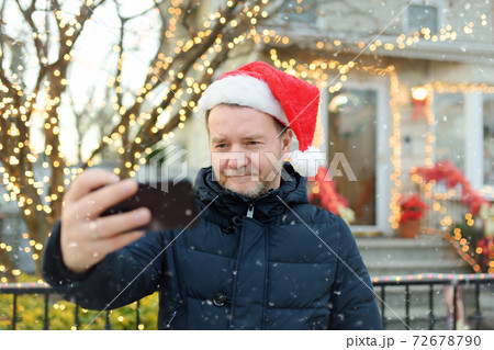 Mature man wearing Santa hat making selfie near his decorated home. Mature man wearing Santa hat making selfie near his decorated home. 72678790