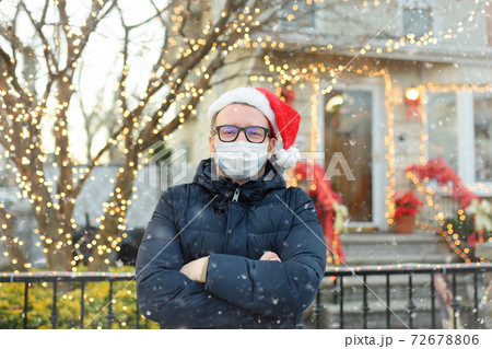 Mature man wearing face mask and Santa hat stand near his decorated home. Mature man wearing face mask and Santa hat stand near his decorated home. 72678806