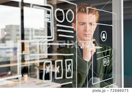 Young pensive male software developer looking at sketches of mobile application Young pensive male software developer looking at sketches of mobile application 72686075
