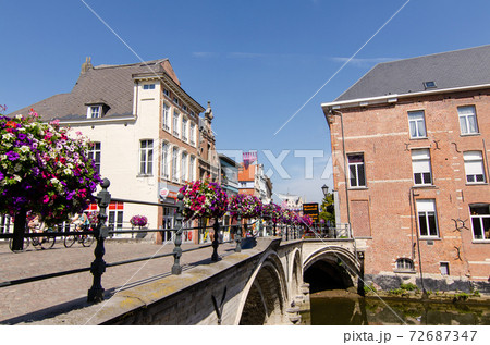 Mechelen, Belgium, August 2019. In the historic center, the bridges are decorated with brightly colored flower boxes. People walk along the sidewalk, beautiful sunny summer day. Mechelen, Belgium, August 2019. In the historic center, the bridges are decorated with brightly colored flower boxes. People walk along the sidewalk, beautiful sunny summer day. 72687347