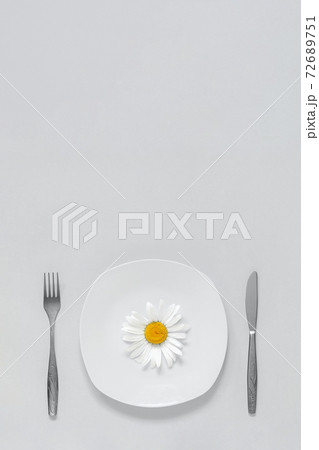 One chamomile flower on plate, cutlery fork and knife on grey background Concept vegetarian healthy eating diet or anorexia. Trendy colors 2021 One chamomile flower on plate, cutlery fork and knife on grey background Concept vegetarian healthy eating diet or anorexia. Trendy colors 2021 72689751