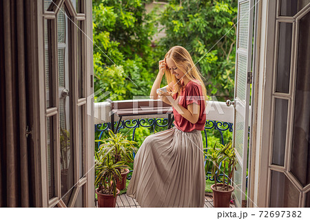 A ginger-haired woman stands on a heritage-style balcony enjoying her morning coffee. A woman in a hotel in Europe or Asia as tourism recovers from a pandemic. Tourism has recovered thanks to 72697382