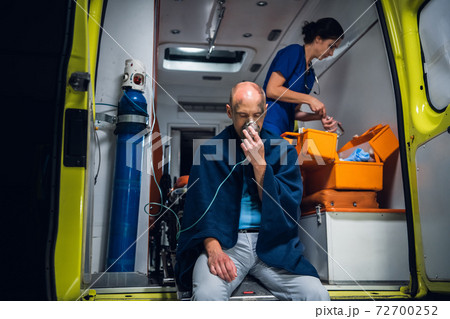 Injured man in a blanket breathing through an oxygen mask in an ambulance car, nurse packing medical equipment in the background Injured man in a blanket breathing through an oxygen mask in an ambulance car, nurse packing medical equipment in the background 72700252