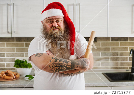 An overweigh man in Santa Claus hat with crossed tattooed arms and a rolling pin on his hand looking at camera thereby showing his emotional state An overweigh man in Santa Claus hat with crossed tattooed arms and a rolling pin on his hand looking at camera thereby showing his emotional state 72715377