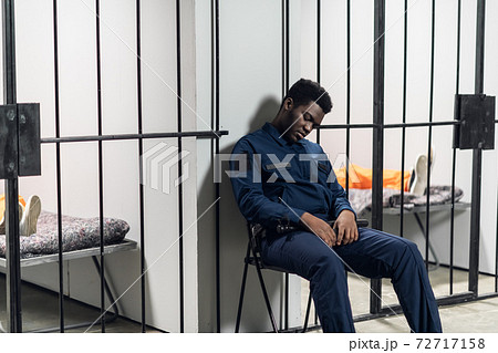 A strong black prison guard guards the cells with prisoners in the prison corridor A strong black prison guard guards the cells with prisoners in the prison corridor 72717158