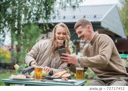 The husband shows his wife the phone screen with something funny. They smile and have a good mood, spending time together on a picnic with barbecue and beer. The husband shows his wife the phone screen with something funny. They smile and have a good mood, spending time together on a picnic with barbecue and beer. 72717452