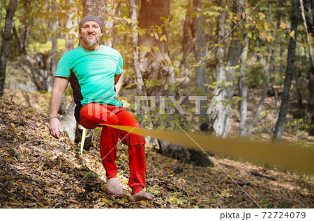 A bearded man in age balances while sitting on a taut slackline in the autumn forest. Outdoor Leisure A bearded man in age balances while sitting on a taut slackline in the autumn forest. Outdoor Leisure 72724079