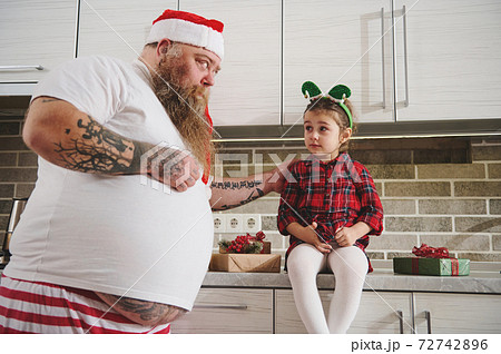 Dad in Santa hat and tattooed arms soothes upset daughter with elf hoop on head.Human Relationship concept. Daughter sitting on a kitchen table.Christmas Gift on the table lie Dad in Santa hat and tattooed arms soothes upset daughter with elf hoop on head.Human Relationship concept. Daughter sitting on a kitchen table.Christmas Gift on the table lie 72742896