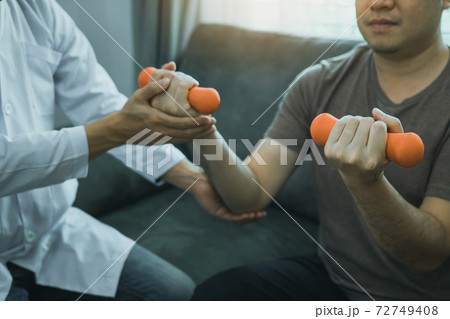 Asian physiotherapist helping a patient lifting dumbbells work through his recovery with weights in clinic room. 72749408