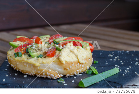 Bagel with hummus, avocado slices, spices, green onion, tomatoes cherry and sesame seeds on the black stone desk. Close up. Bagel with hummus, avocado slices, spices, green onion, tomatoes cherry and sesame seeds on the black stone desk. Close up. 72755679