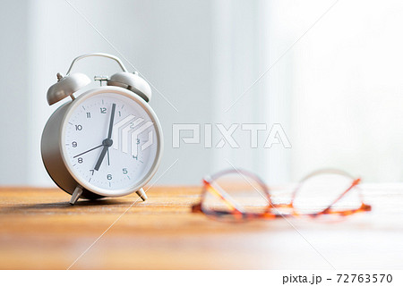 窓辺の机の上に置かれた朝日に照らされる目覚まし時計とメガネ 72763570