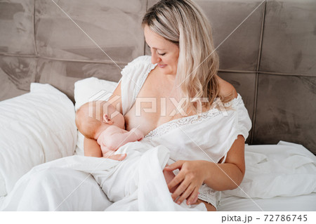 woman is sitting with a newborn in a white bed feeding breast milk. 72786745