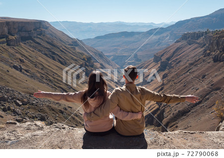 Excited couple sits on rocky cliff edge against canyon Excited couple sits on rocky cliff edge against canyon 72790068