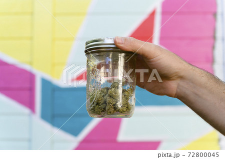 Close-up of mans hand holding jar with cannabis buds in front of colorful mural Close-up of mans hand holding jar with cannabis buds in front of colorful mural 72800045