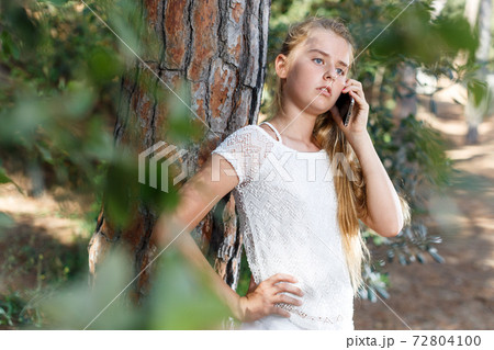 Serious teenage girl talking by phone near tree in green park 72804100