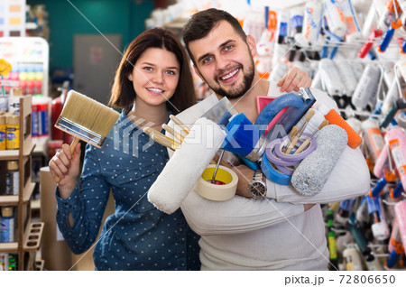 Young cheerful couple demonstrating tools for house renovating in paint store Young cheerful couple demonstrating tools for house renovating in paint store 72806650