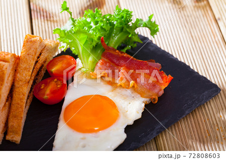 Cooked eggs with bacon, toasted bread, tomatoes and lettuce at plate on table Cooked eggs with bacon, toasted bread, tomatoes and lettuce at plate on table 72808603