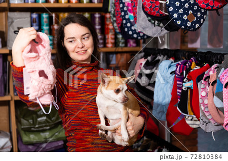 Positive woman holding dog and choosing clothes in petshop 72810384