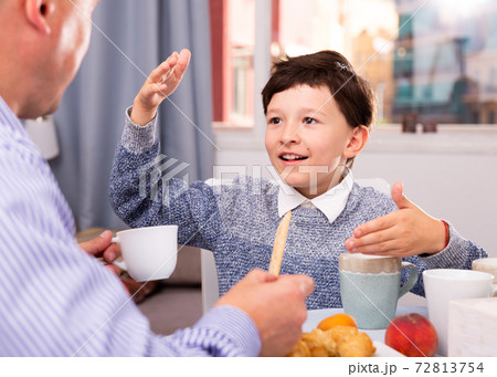 Son talking with father at table with tea during breakfast indoors 72813754