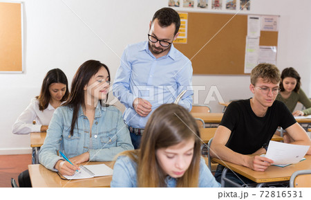 Smiling schoolgirl is writing answer the question on paper and teacher explaining it 72816531