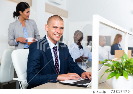 Cheerful businessman sitting at workplace in open plan office Cheerful businessman sitting at workplace in open plan office 72817479
