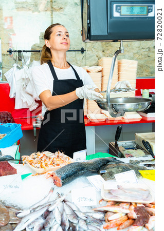 Confident female fishmonger working behind store counter, weighing fresh anchovies on scales 72820021