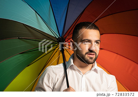 Bearded guy hold colorful umbrella. A nice guy with a stylish beard and white shirt stands with a rainbow umbrella behind his back. Handsome guy in support of LGBT society 72833572