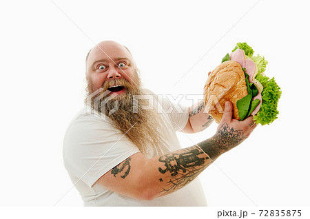 A bearded man with tattooed hands holding a huge burger in his hands and smiling with joy showing it to the camera A bearded man with tattooed hands holding a huge burger in his hands and smiling with joy showing it to the camera 72835875