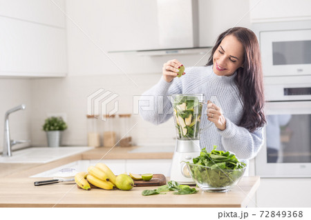 Woman standing in a kitchen squeezing lime into a blender full of fruit and vegetable 72849368