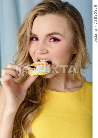 European young blonde woman with wavy hair  bites a multicolored cookie in her hand, 72850559