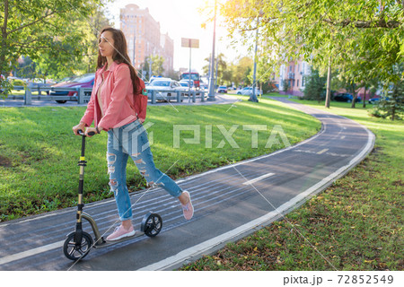 girl in summer in city rides a scooter, in casual clothes jeans and a pink jacket, free space for a copy of text. Background road green grass and trees, motorway highway and cars. 72852549