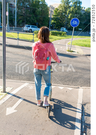 woman in city in summer stands at crossroads with a scooter. We are waiting for traffic signal to cross road. Jeans jacket and backpack behind back. 72852550