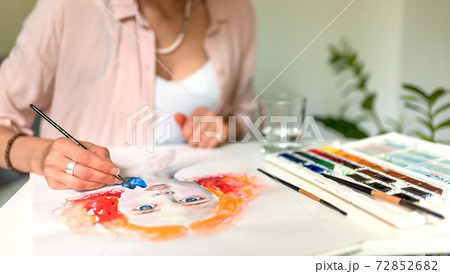 Close-up of female hands are painting a picture, a pink shirt at the artist, a portrait of a drawing girl, orange hair, a palette with flowers, a glass of water, brushes. Close-up of female hands are painting a picture, a pink shirt at the artist, a portrait of a drawing girl, orange hair, a palette with flowers, a glass of water, brushes. 72852682