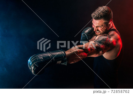 Bearded tattooed sportsman muay thai boxer in black undershirt and boxing gloves fighting on dark background with smoke. 72858702
