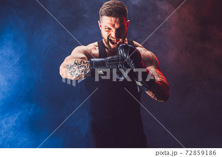 Bearded tattooed sportsman muay thai boxer in black undershirt and boxing gloves fighting on dark background with smoke. 72859186
