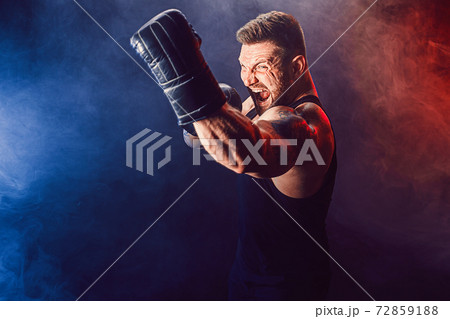 Bearded tattooed sportsman muay thai boxer in black undershirt and boxing gloves fighting on dark background with smoke. 72859188