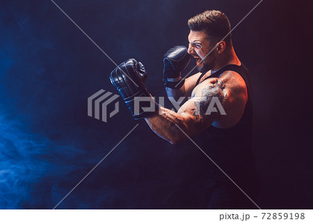 Bearded tattooed sportsman muay thai boxer in black undershirt and boxing gloves fighting on dark background with smoke. Bearded tattooed sportsman muay thai boxer in black undershirt and boxing gloves fighting on dark background with smoke. 72859198