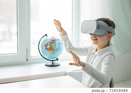 A child with a virtual reality headset is sitting at the table indoors at home. A girl exploring the world of virtual reality through video and games. A child with a virtual reality headset is sitting at the table indoors at home. A girl exploring the world of virtual reality through video and games. 72859489