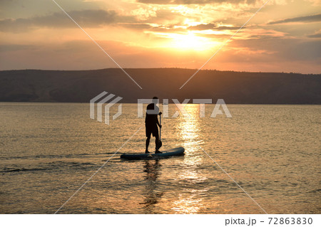 Man sail on a SUP board in a large lake during sunrise. Stand up paddle boarding active recreation in nature Man sail on a SUP board in a large lake during sunrise. Stand up paddle boarding active recreation in nature 72863830