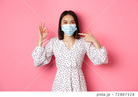 Coronavirus, social distancing and lifestyle concept. Cute asian woman pointing at face mask, showing okay sign, support using preventive measures from covid-19, pink background Coronavirus, social distancing and lifestyle concept. Cute asian woman pointing at face mask, showing okay sign, support using preventive measures from covid-19, pink background 72867328