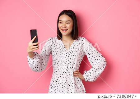 Beautiful asian girl using photo filters app and taking selfie on smartphone, posing in cute dress against pink background Beautiful asian girl using photo filters app and taking selfie on smartphone, posing in cute dress against pink background 72868477