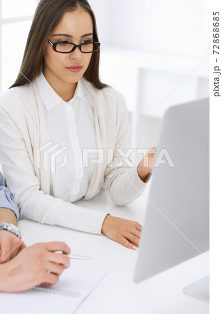 Young woman sitting at the desk with computer in white colored office. Looks like student girl or business lady communicating with casual dressed man Young woman sitting at the desk with computer in white colored office. Looks like student girl or business lady communicating with casual dressed man 72868685