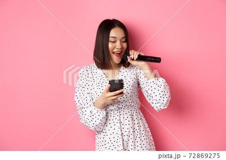 Cute asian woman reading lyrics in smartphone, singing with microphone, standing in trendy dress over pink background 72869275
